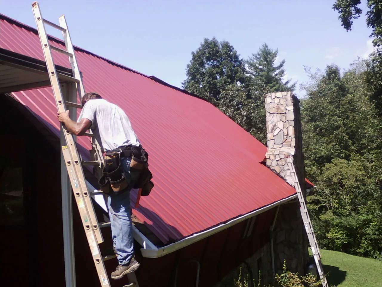 Roofer on a ladder beside a red metal roof and stone chimney — Soffit & Fascia Repair by Roofing Brothers in Negley
